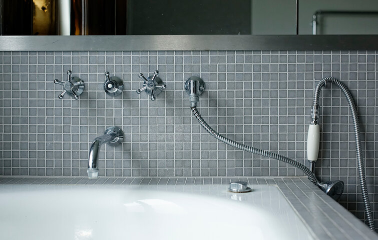 Udsnit af badeværelse med hvidt badekar og grå mosaikfliser på væggen.